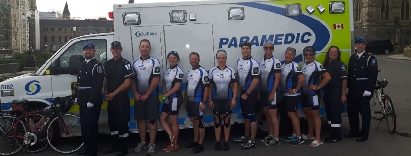 A group of six male and six female paramedics with bikes lined up in front of an ambulance.
