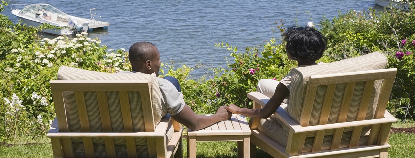 A couple sitting in wooden chairs by the lake holding hands.