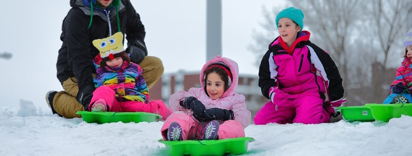 Two people pushing two little girls on green sleds down a hill.