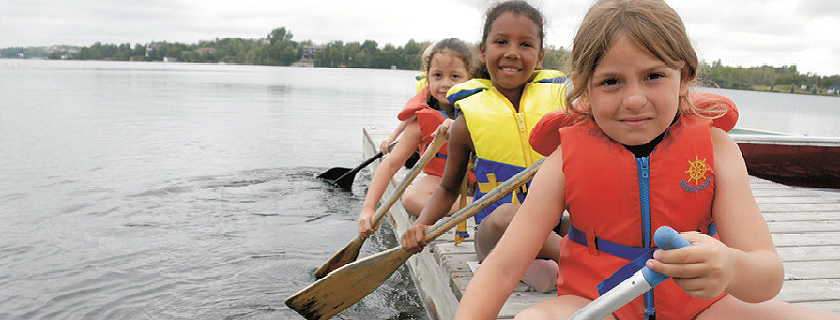 Children sitting on the side of a dock holding canoe paddles.