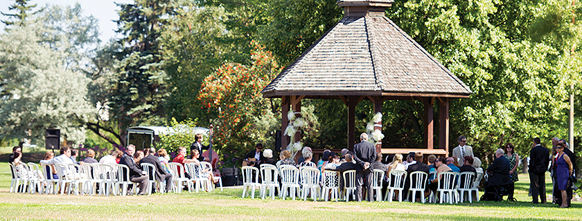 An outdoor wedding at a gazebo with white chairs and an audience.
