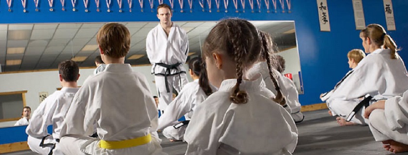 Nine children sitting down in white karate uniforms watching their instructor.