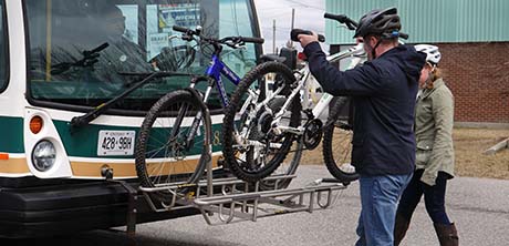 Male cyclist lifting one of two bicycles onto the rack on the front of a transit bus.